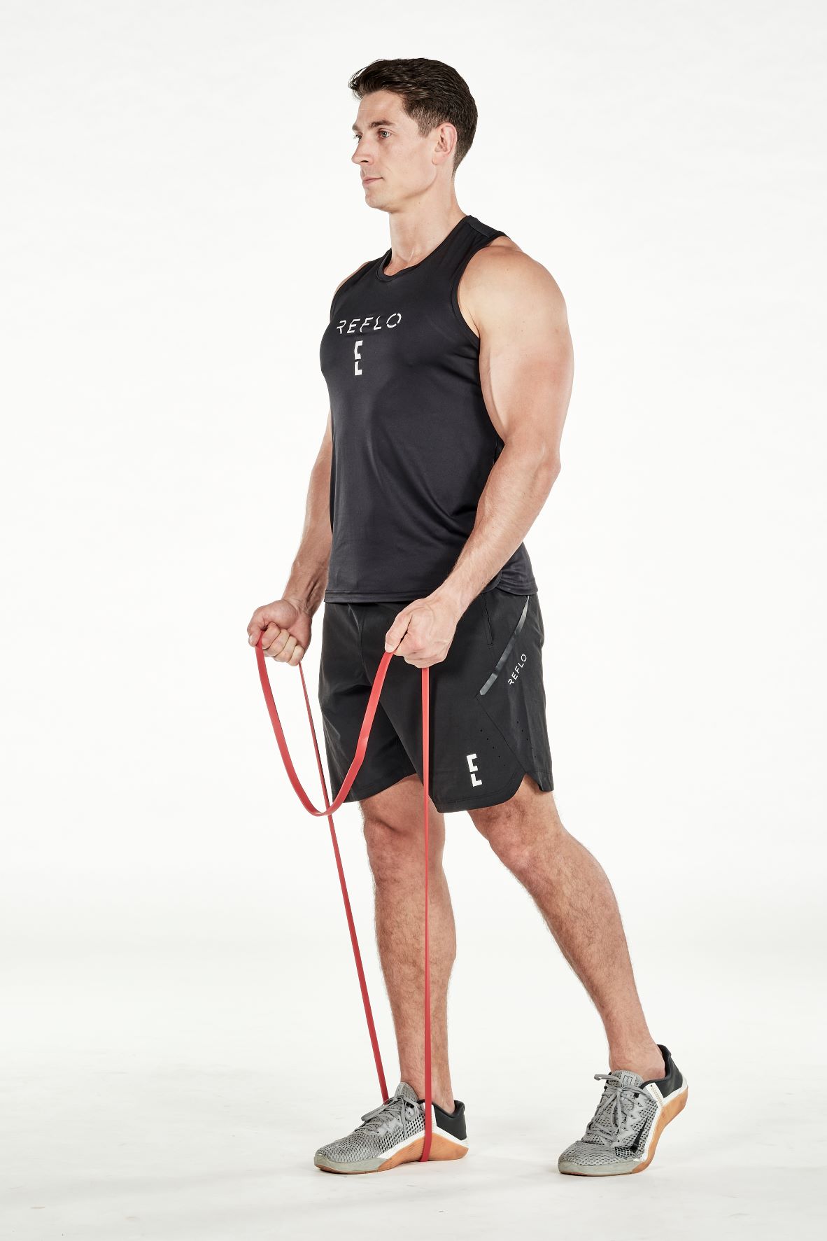 man demonstrating step one of resistance band biceps curl; standing up straight, he is holding a resistance band that is wrapped under one foot; he wears a black fitness vest, black shorts and trainers