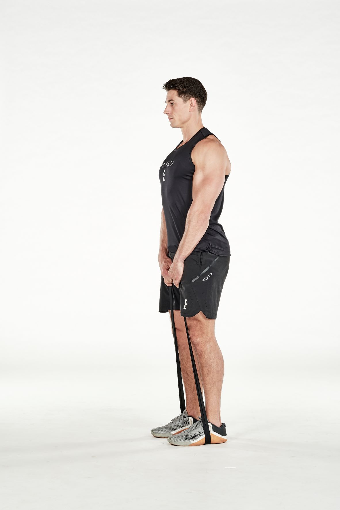 man demonstrating step two of resistance band deadlift; he is standing up straight, holding a resistance band that is wrapped around his feet; he wears a black fitness vest, black shorts and trainers