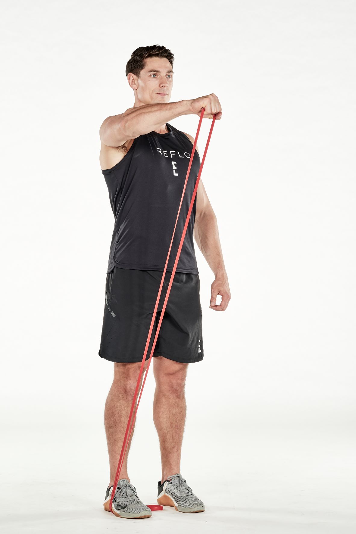 man demonstrating step two of resistance band front raise; standing up straight, he is holding a resistance band that is wrapped under one foot in one hand; keeping the arm straight, he lifts and extends his arm up until it is level with his shoulder; he wears a black fitness vest, black shorts and trainers
