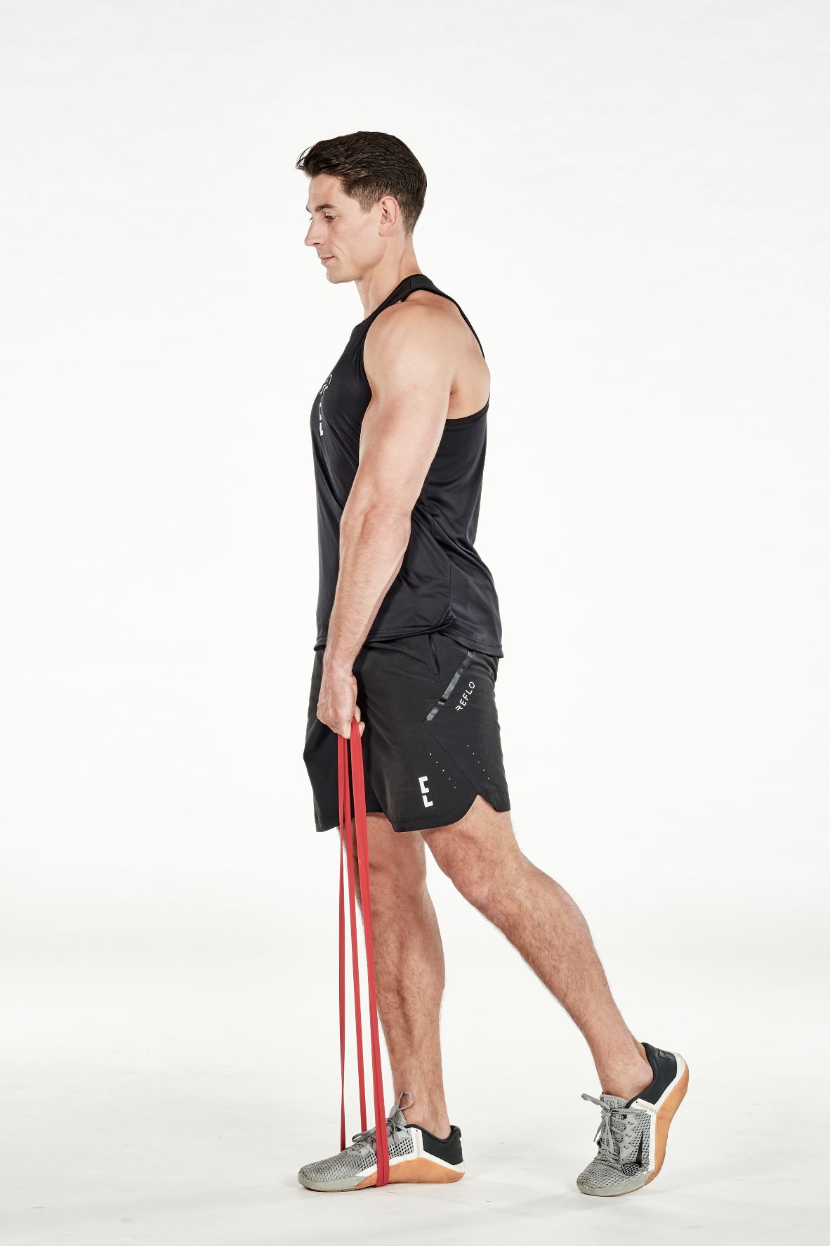man demonstrating step two of resistance band single-leg romanian deadlift; he stands straight, holding a resistance band which is being held under his foot; his other leg is relaxed and positioned behind him; he wears a black fitness vest, black shorts and trainers
