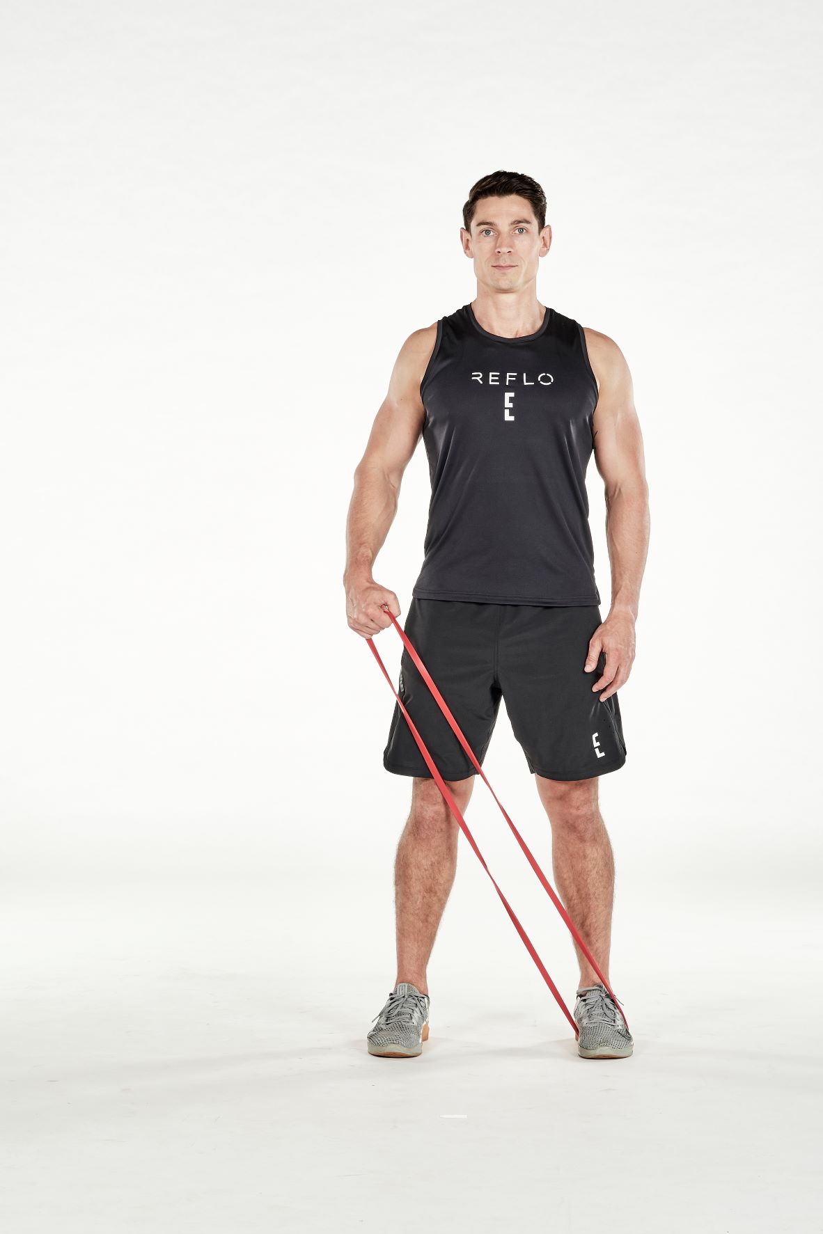 man demonstrating step one of resistance band lateral raise; standing up straight, a resistance band is held under one foot; he holds the resistance band at hip height with the opposite hand; he wears a black fitness vest, black shorts and trainers