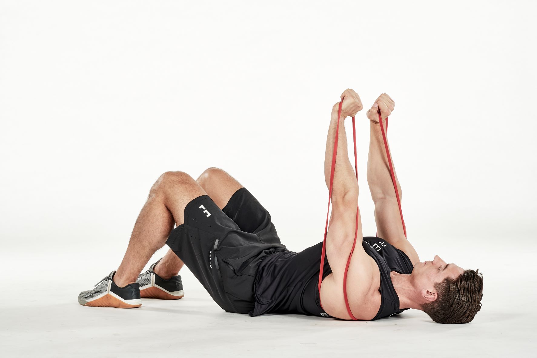 man demonstrating step two of lying chest press; laying on his back with bent knees, the resistance band is passed under his back; holding one end of the resistance band in each hand, he extends his arms up towards the ceiling; he wears a black fitness vest, black shorts and trainers