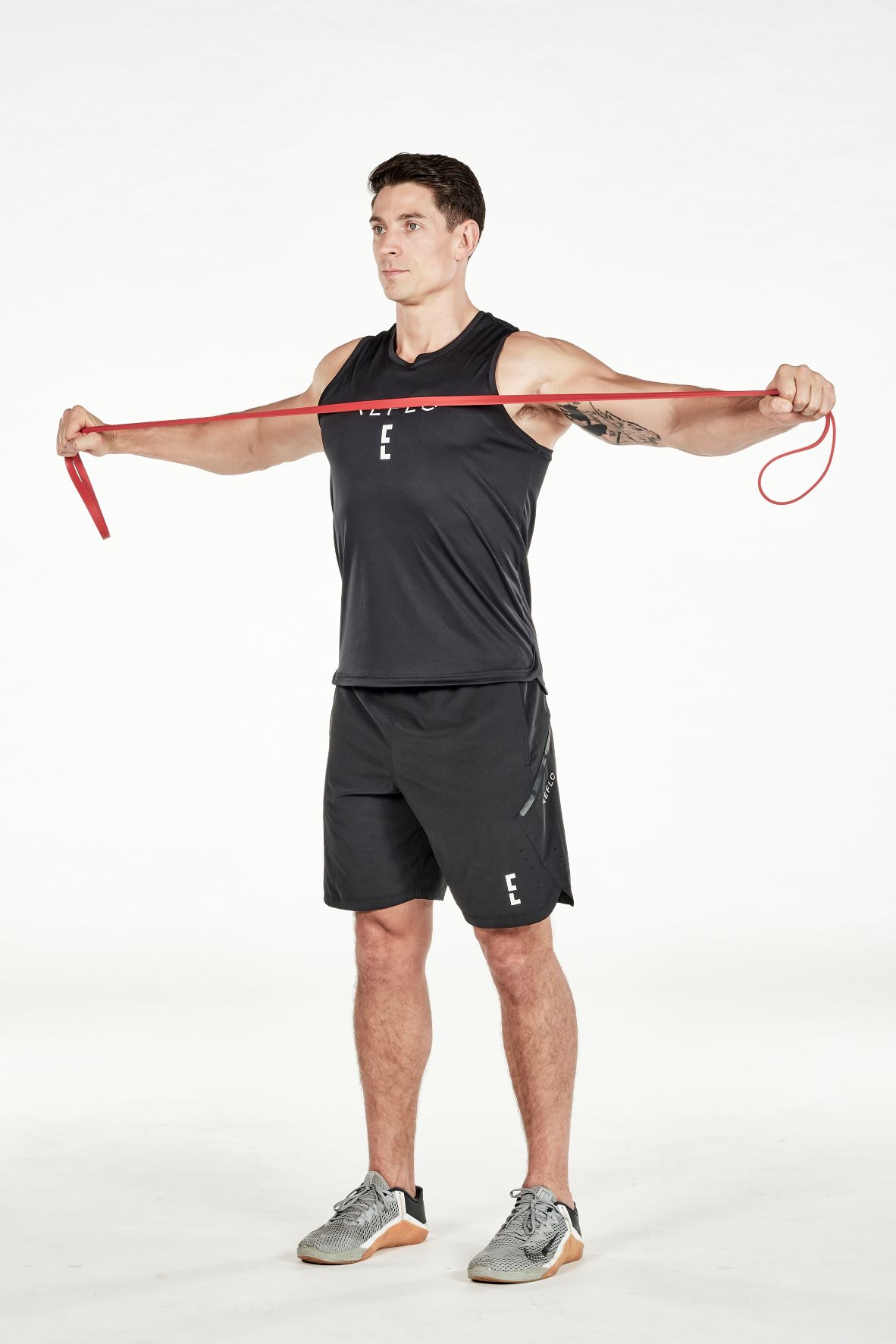 man demonstrating step two of reverse fly; standing up straight, he holds a resistance band in both hands, he stretches his arms outwards as far as possible; he wears a black fitness vest, black shorts and trainers