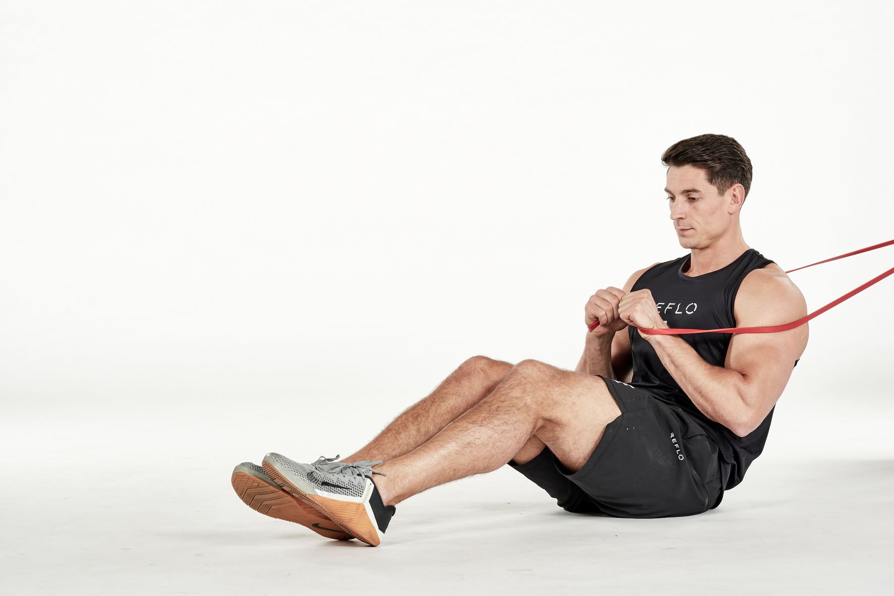 man demonstrating step one of russian twist; sitting down with knees bent, he holds a secured resistance band in his hands in front of his chest; he wears a black fitness vest, black shorts and trainers
