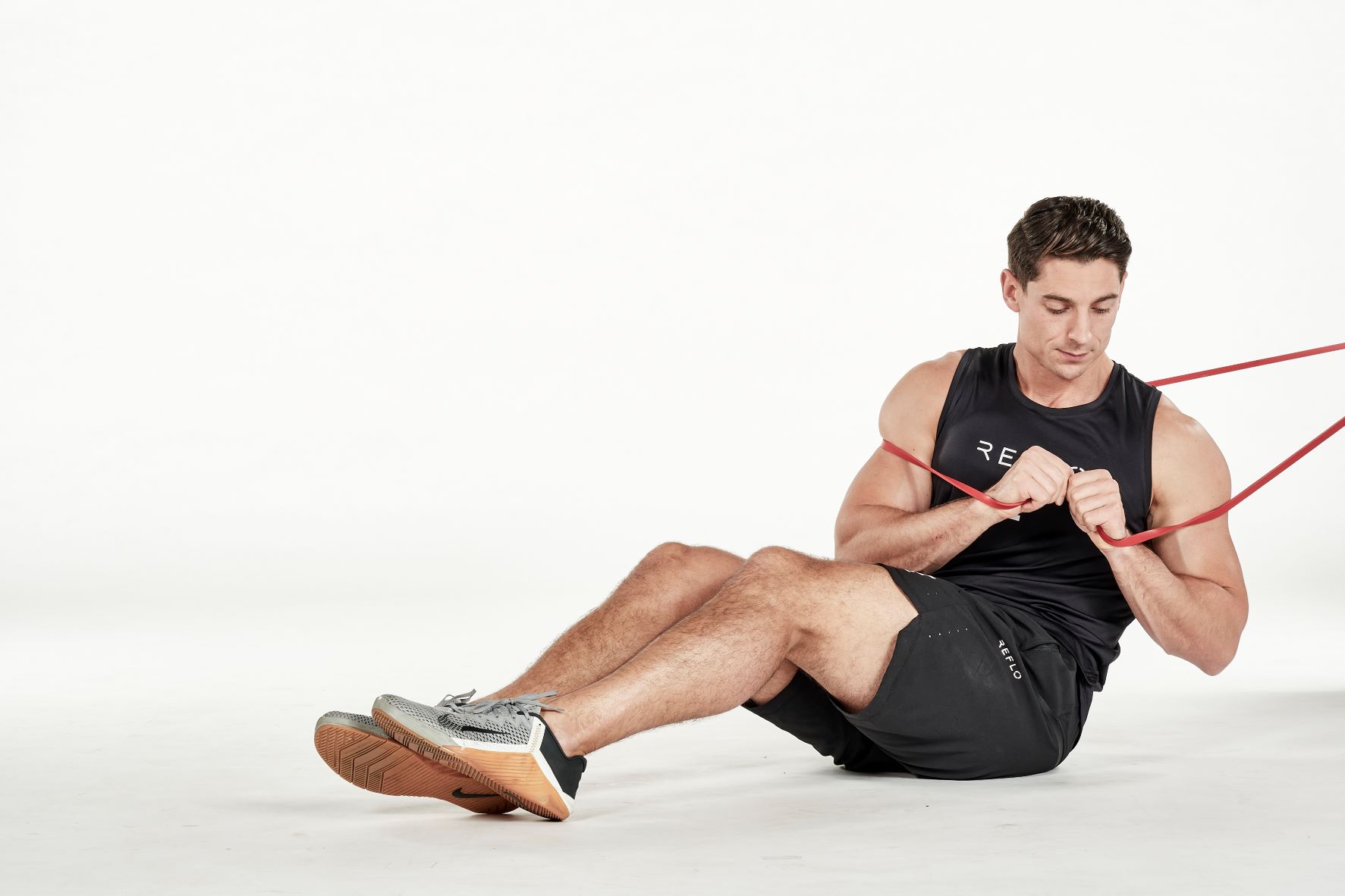 man demonstrating step two of russian twist; sitting down with knees bent, he holds a secured resistance band in his hands in front of his chest; he turns his body to one side; he wears a black fitness vest, black shorts and trainers