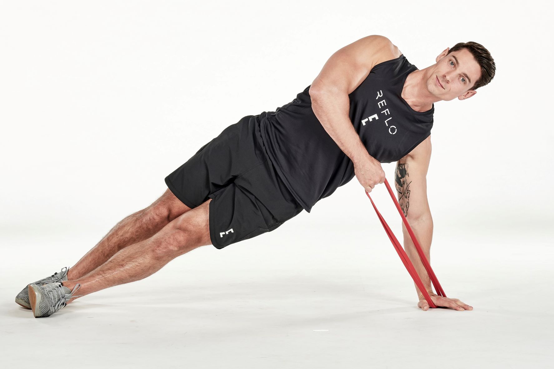 man demonstrating step one of side plank high pull; in a side plank position, his top hand holds a resistance band that is secured under his bottom hand on the floor; he wears a black fitness vest, black shorts and trainers