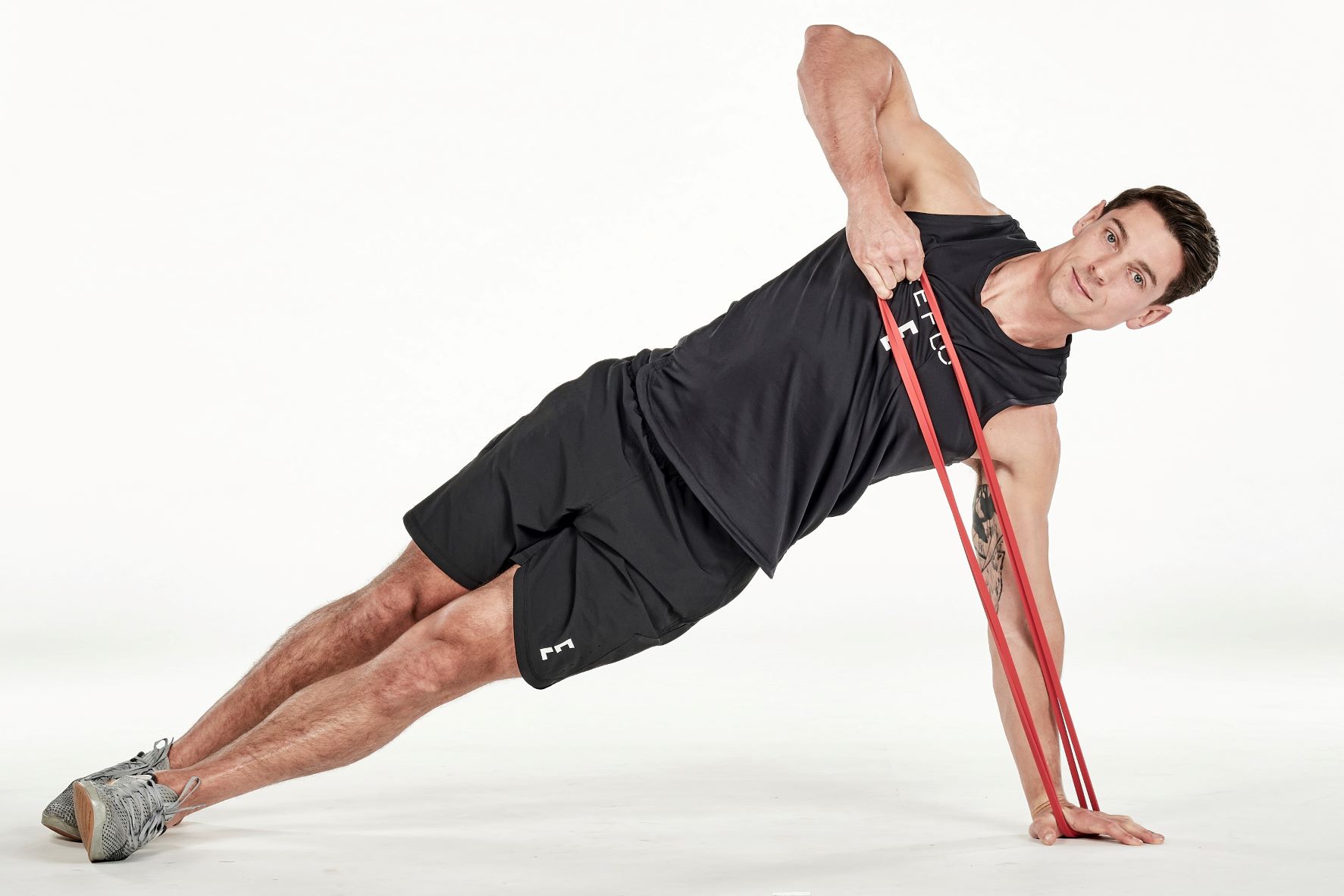 man demonstrating step two of side plank high pull; in a side plank position, his top hand holds a resistance band that is secured under his bottom hand on the floor; his top arm is bent as he pulls the band upwards; he wears a black fitness vest, black shorts and trainers