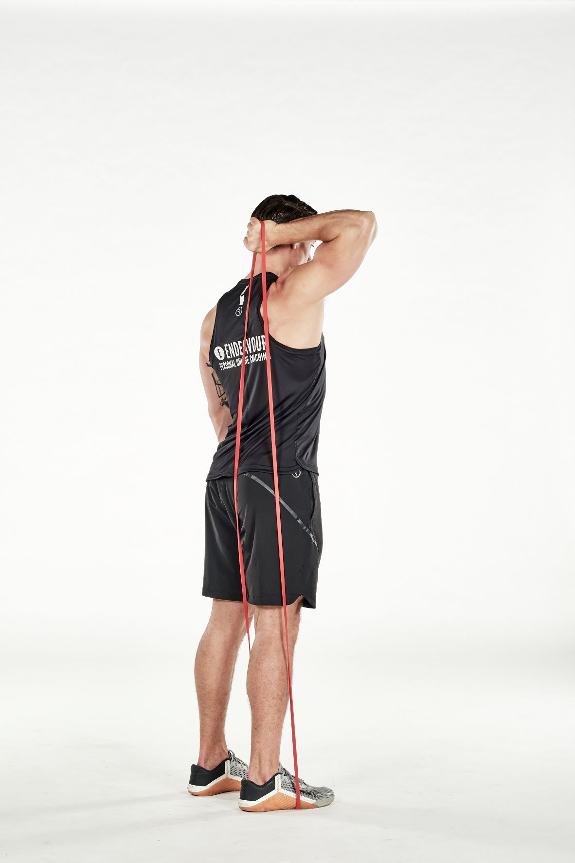 man demonstrating step one of single arm triceps extension; standing tall, his arm reaches behind his head to hold a resistance band that's held under one foot; his arm is bent; he wears a black fitness vest, black shorts and trainers