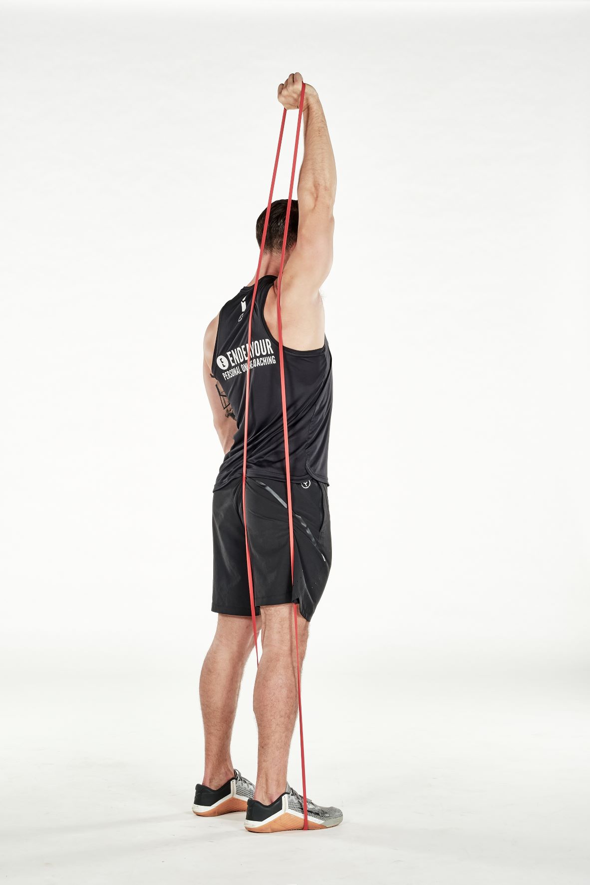 man demonstrating step two of single arm triceps extension; standing tall, his arm reaches behind his head to hold a resistance band that's held under one foot; his arm straightens as he pulls the band and reaches up; he wears a black fitness vest, black shorts and trainers