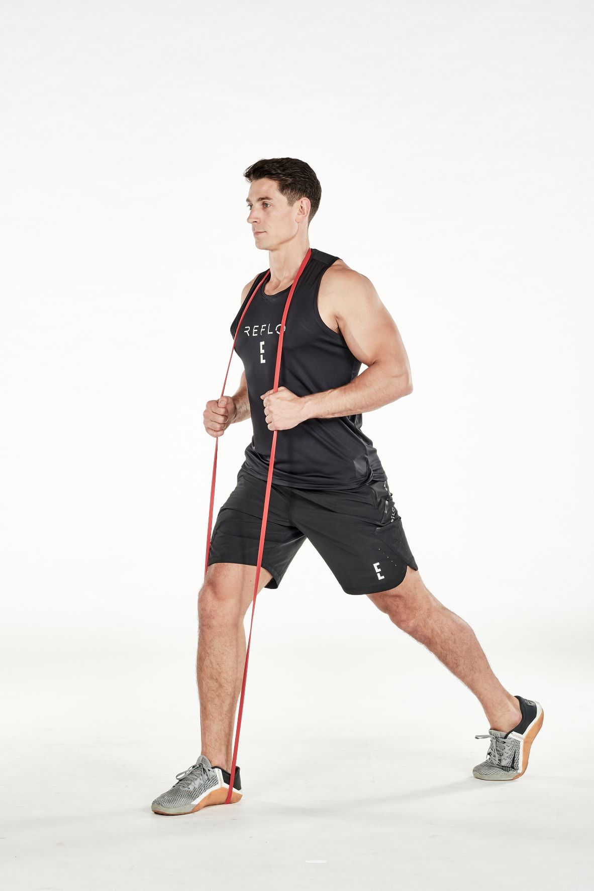 man demonstrating step one of split squat; legs apart, he holds a resistance band under his front foot and around the back of his neck; his hands support the band in the middle; his arm is bent; he wears a black fitness vest, black shorts and trainers
