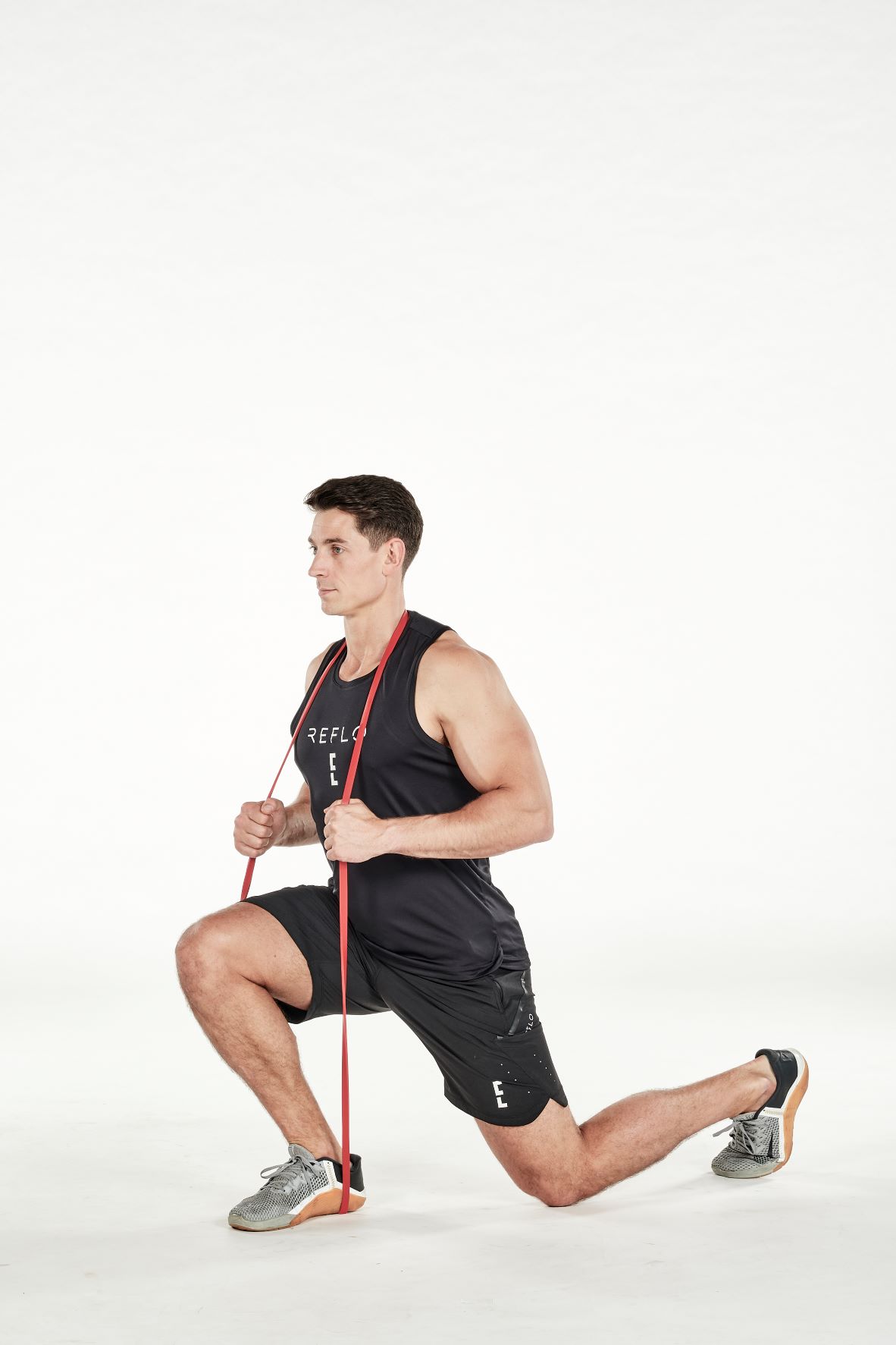 man demonstrating step two of split squat; bending down into a lunge position, he holds a resistance band under his front foot and around the back of his neck; his hands support the band in the middle; his arm is bent; he wears a black fitness vest, black shorts and trainers