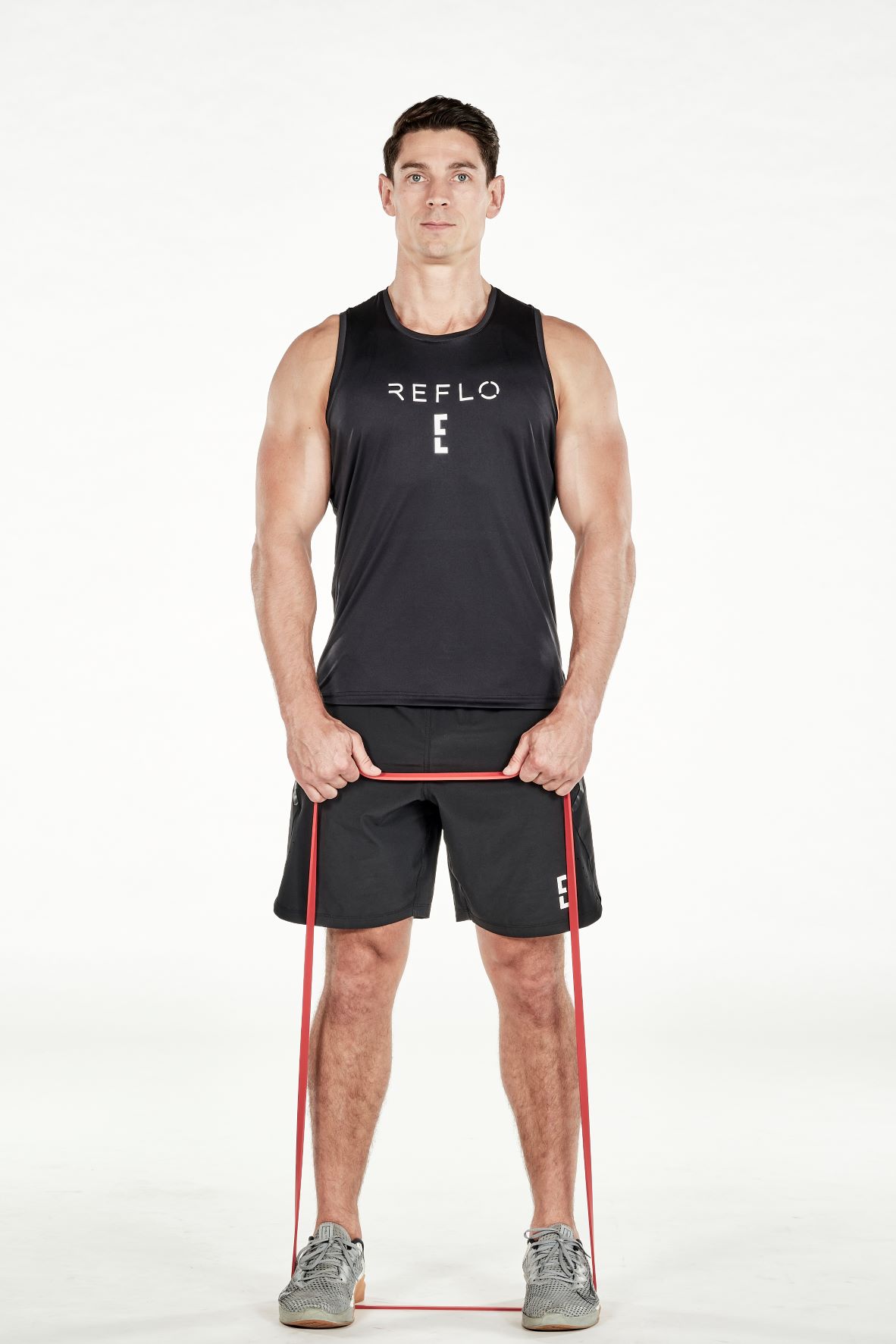 man demonstrating step one of upright row; he stands upright, both hands holding a resistance band that is secured under his feet; he wears a black fitness vest, black shorts and trainers