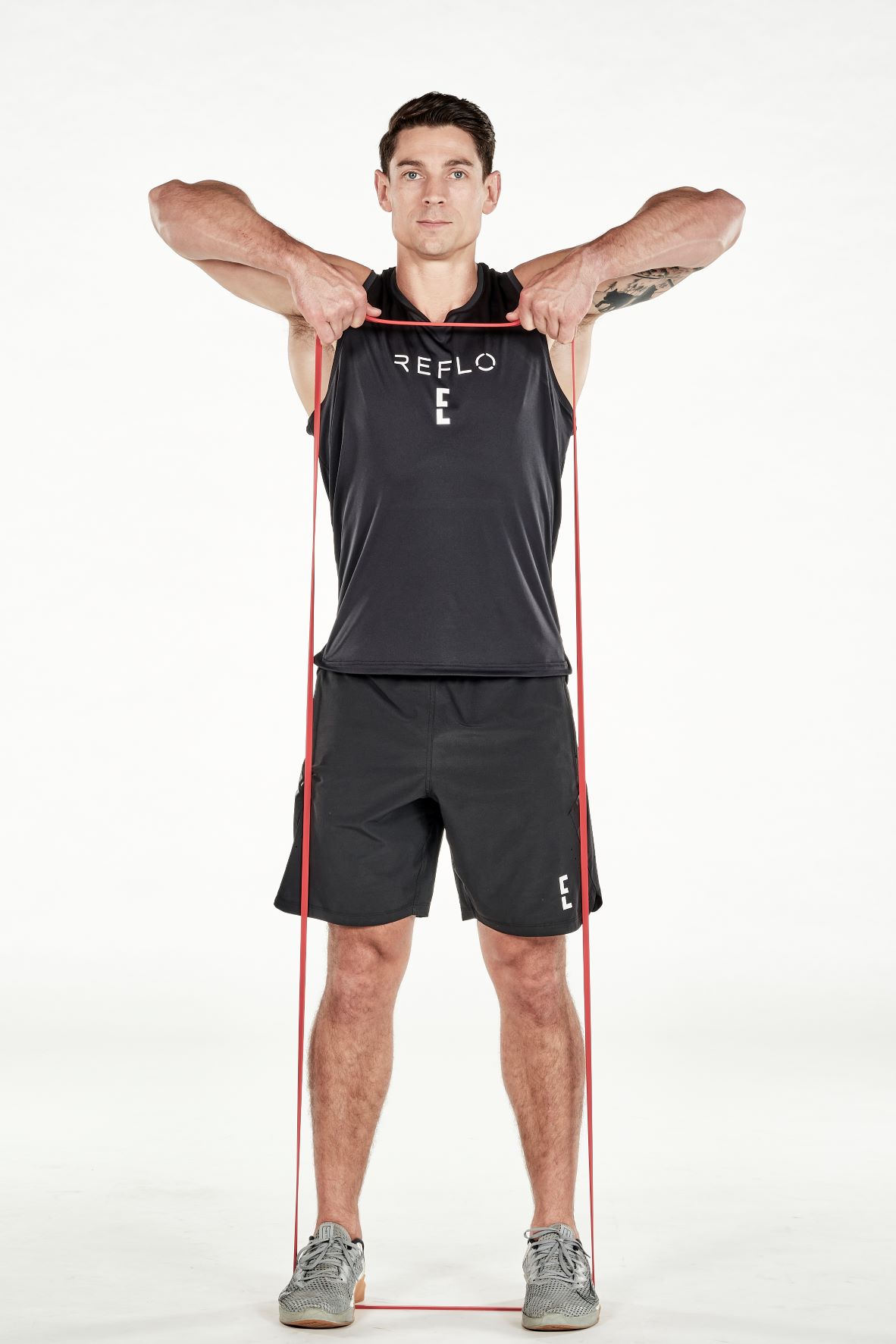 man demonstrating step two of upright row; he stands upright, both hands holding a resistance band that is secured under his feet; he pulls up on the band until level with his shoulders; he wears a black fitness vest, black shorts and trainers