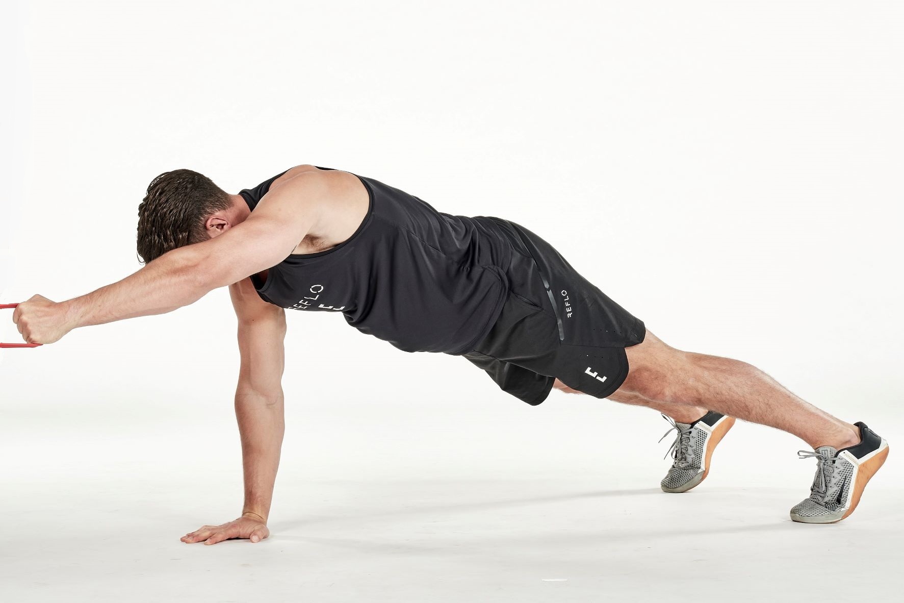 man demonstrating step one of plank row; in a plank position, one arm reaches out holding a secured resistance band; he wears a black fitness vest, black shorts and trainers