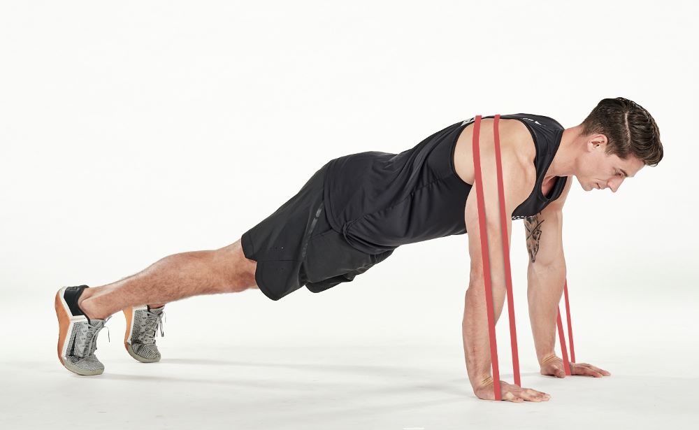 man demonstrating step one of press up; in a plank position, the resistance band passes over his back to be held by each hand on the floor; he wears a black fitness vest, black shorts and trainers