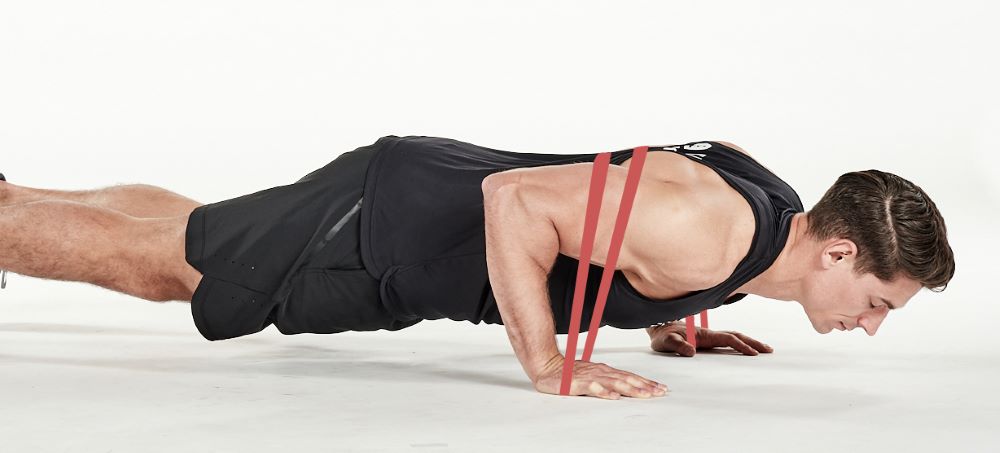 man demonstrating step two of press up; with the resistance band passing over his back to be held by each hand on the floor, he bends his arms to lower his body towards the floor; he wears a black fitness vest, black shorts and trainers