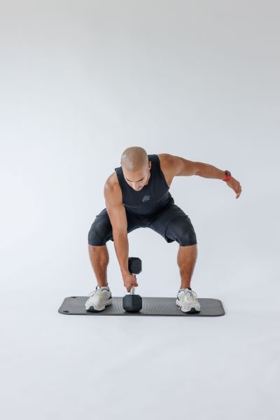 demonstration of dumbbell single arm snatch, step 1: Stand with your feet shoulder width apart, holding a dumbbell in one hand. Push your hips back and lower yourself into a deep squat.   Allow the dumbbell to hang down to touch the floor directly beneath your chest keeping that arm straight. Your back should also be straight and aim to keep the chest elevated. 