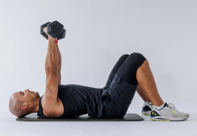 man demonstrates floor chest press: he lays on his back, knees bent, pressing two dumbbells up towards the sky