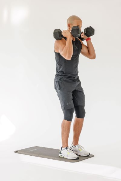 man demonstrating reverse lunge press step 1: he stands upright with two dumbbells held at his shoulders