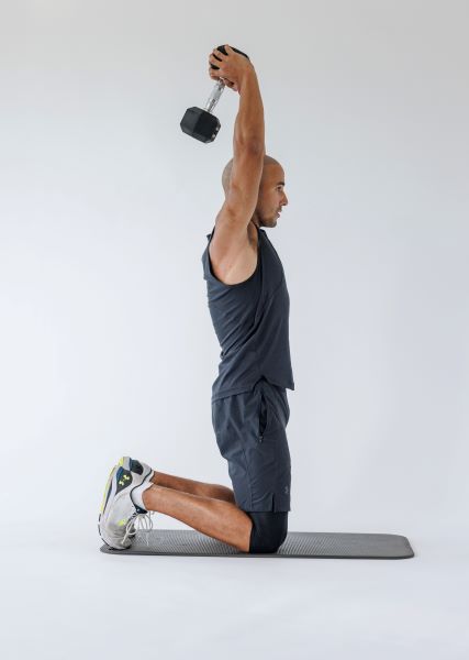 man demonstrating tricep overhead extension: kneeling, he presses a dumbbell above his head using both hands