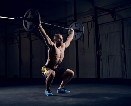 Man with bare chest squatting to lift a barbell in a gym - get stronger by training less