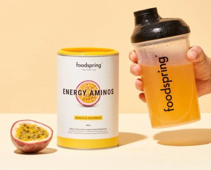 Product shot of a tub of Foodspring Energy Amino powder, a hand with a drink bottle and half a passionfruit