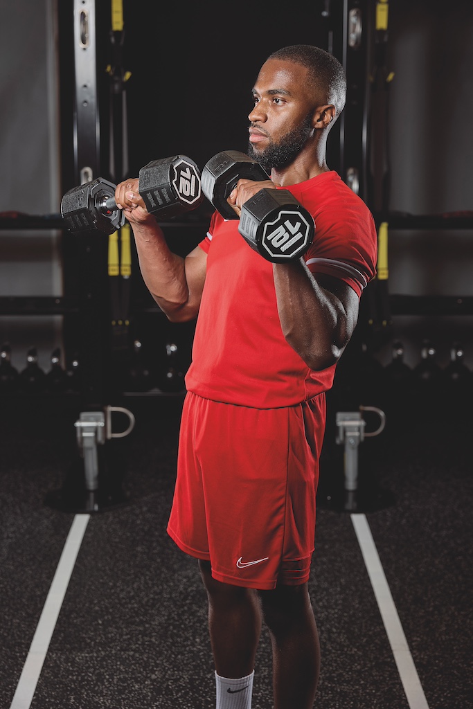 man in red top and red shorts doing a dumbbell reverse-grip biceps curl as part of a full-body circuit