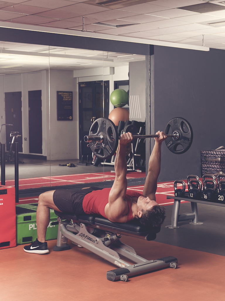 man in red vest and black shorts performing EZ-bar triceps extension in the gym