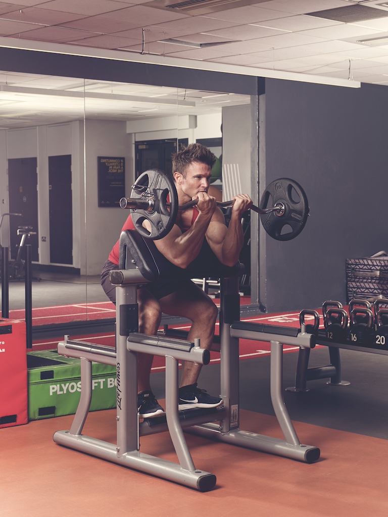 man in red vest and black shorts performing EZ-bar preacher curl in the gym – one of the best barbell arms exercises