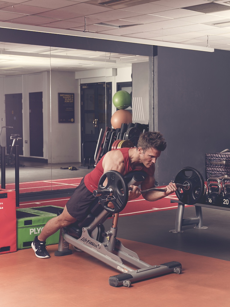 man in red vest and black shorts performing EZ-bar prone incline curl in the gym – one of the best barbell arms exercises