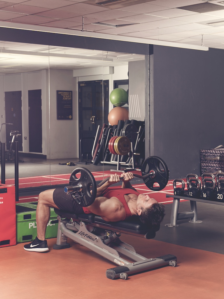 man in red vest and black shorts performing EZ-bar triceps press in the gym