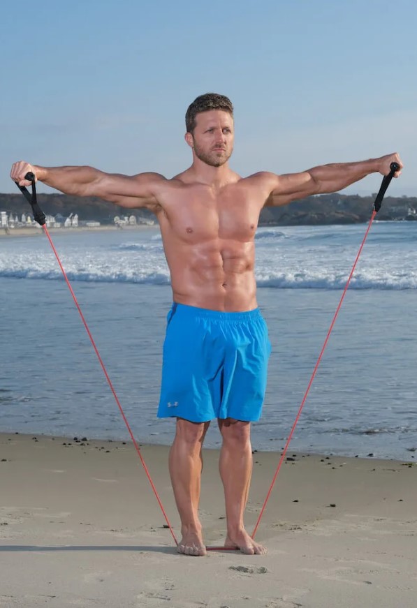 Man performing a resistance band lateral raise on a beach