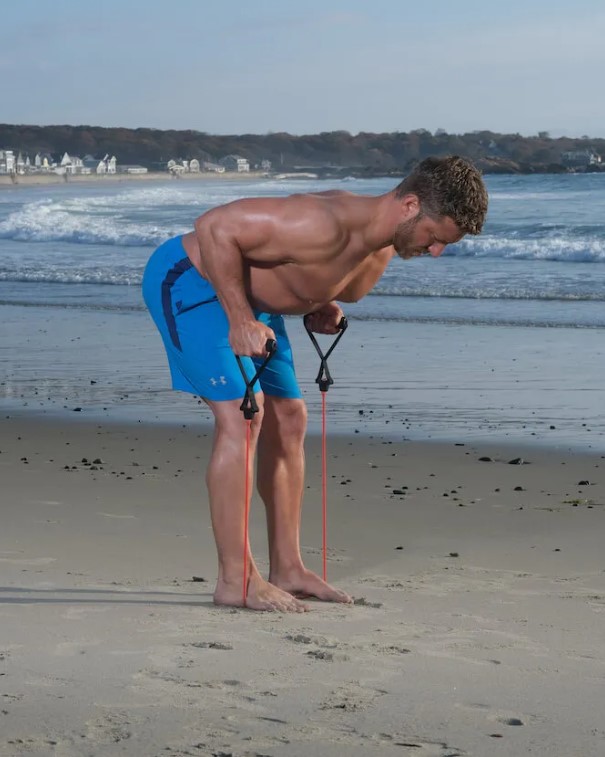 Man performing a resistance band bent-over row on a beach