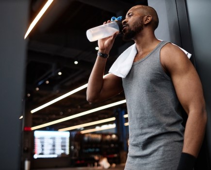 A man leaning against a gym wall drinking