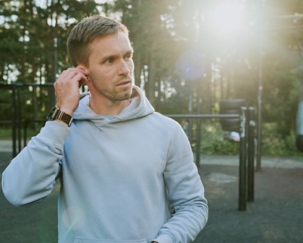 Caucasian young adult man adjusting wireless earbud while standing outdoors in park with fitness equipment, sunlight shining through trees in background, wearing casual hoodie