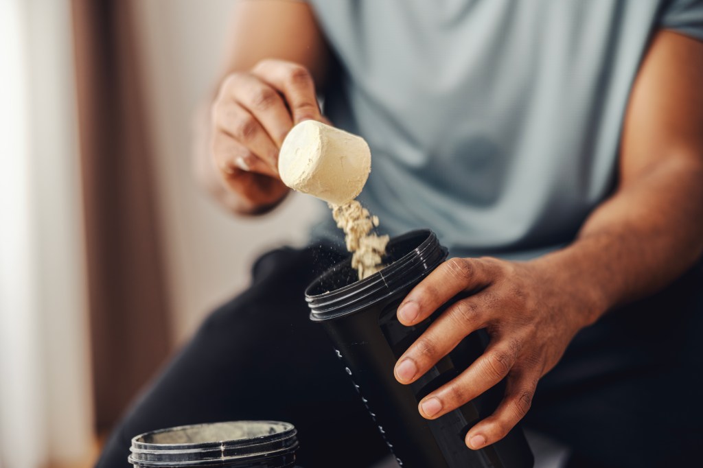Adult man's hands scooping protein powder from a container into a black shaker bottle, preparing a nutritional supplement drink for workout recovery and muscle building