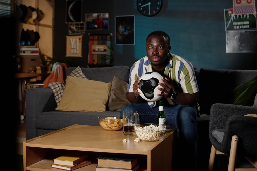 Young African American guy holding ball sitting on sofa in living room late in evening watching soccer match on TV