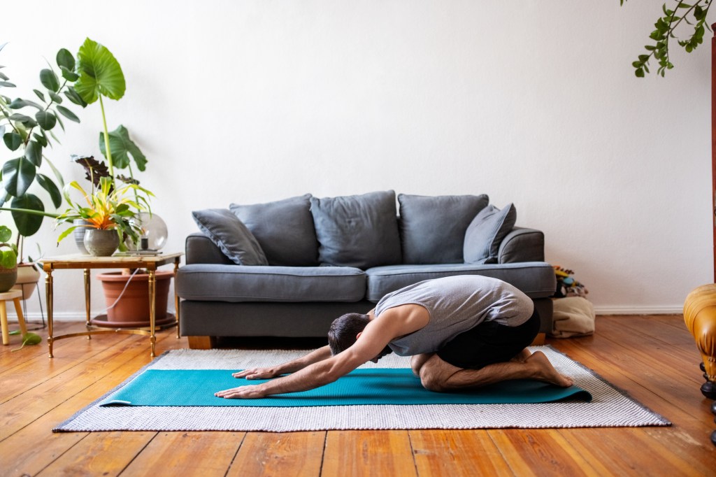 Side view of a mid adult male working out at home. Man doing childs pose yoga on exercise mat indoors.