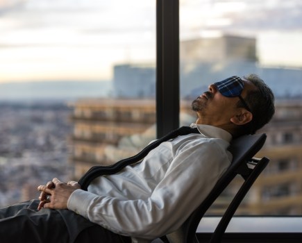 A man reclining in a chair from right to left in an office building during the working day wearing an eye mask to help him de-stress