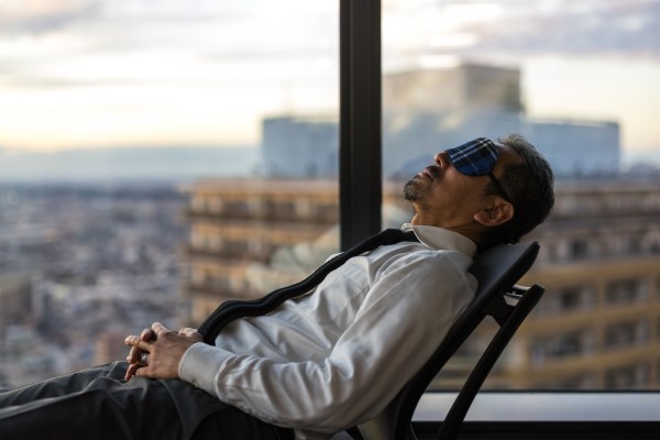 A man reclining in a chair from right to left in an office building during the working day wearing an eye mask to help him de-stress